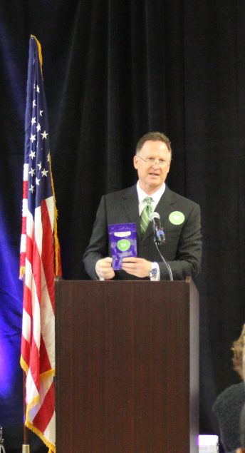 A man standing behind a wooden podium with an American flag on the left side. He is wearing a dark suit and tie and is holding a purple book in his hands. He appears to be giving a speech or presentation. The background is black and there is a blue curtain behind him. There are a few people sitting in front of the podium listening attentively to the man.