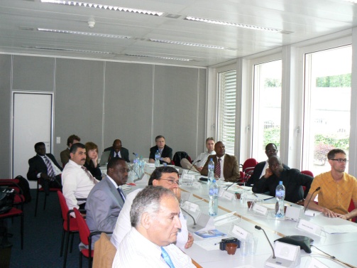 A large conference room with a long table in the center. The table is covered with a white tablecloth and has nameplates water bottles and microphones on it. There are several people sitting at the table all dressed in formal business attire. The room has large windows on the right side allowing natural light to enter. The walls are painted white and there is a door on the left side of the room. The people in the room appear to be engaged in a discussion or meeting.