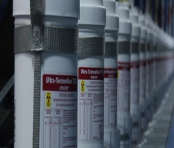 A row of white cylindrical containers with red labels on them. The labels have the words "Ultra-Technicool" written in bold black letters at the top followed by "Ultra Technicool". Below the text there is a yellow warning sign with a red exclamation mark. The containers are arranged in a neat row and appear to be made of plastic or metal. The background is blurred but it seems to be a warehouse or storage area with shelves and other equipment visible.