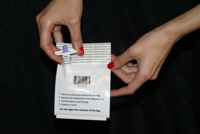 A pair of hands holding a small white packet with a purple label on it. The label has a barcode and some text on it which appears to be instructions on how to use the packet. The packet is open and the person's hands are holding it with their fingers. The background is black and the focus is on the hands and the packet itself.