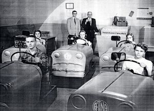 A black and white photograph of a group of children in a room. Each of the five children are sitting behind the wheel of their own toy car. The cars are arranged in a semi-circle around the room with the front of the cars facing towards the left side of the image. The cars have decals reading "ETNA".<br /><br />In the center of the room behind the children are two men wearing a suit and tie standing next to a desk with unidentified equipment. The photograph appears to have been taken in the mid-twentieth century.