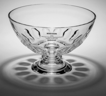 A black and white photograph of a glass bowl. The bowl is made of clear glass and has a round base with a pedestal base. The rim of the bowl is slightly curved and has the word "McKesson" engraved on it in a cursive font. The background is plain white making the bowl the focal point of the image. The image is taken from a slightly elevated angle casting a shadow on the surface below the bowl.