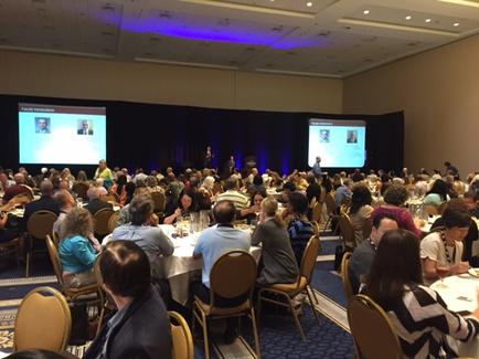 A large conference room with a large group of people sitting at tables facing a stage. The room has a high ceiling with recessed lighting and a carpeted floor. On the stage there are two large screens displaying images of people and text. The people in the room appear to be engaged in a discussion or presentation. The tables are covered with white tablecloths and there are glasses of water on the tables. The overall atmosphere of the room is formal and professional.