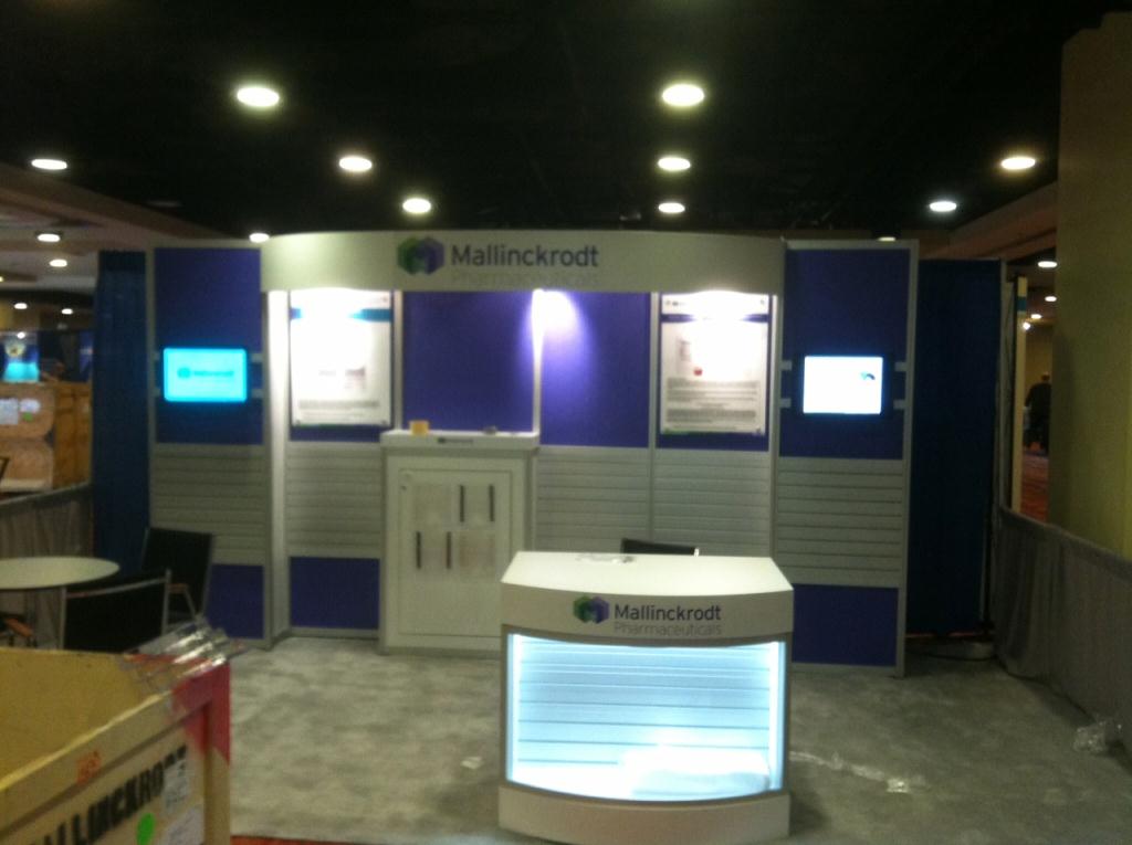 A trade show booth with a blue and white color scheme. The booth has a sign that reads "Mallinckrodt" in bold white letters. There are two large screens on either side of the booth displaying information about the company's products. In front of the screen there is a small white counter with a glass display case. The floor is covered with a gray carpet and there are several other booths and tables in the background. The overall atmosphere of the image is professional and modern.