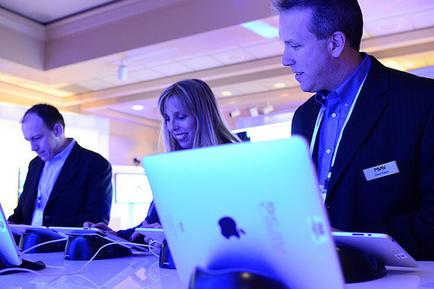 Three people working on laptops in an office setting. There are three people in the image two men and a woman sitting at a desk with their laptops in front of them. The man on the right is wearing a black suit and a name tag while the man in the middle is looking intently at one of the laptops. The woman is smiling and appears to be focused on the screen. The desk is covered with a white tablecloth and there are several other laptops on it. The background is blurred but it seems to be an indoor space with a high ceiling and large windows. The image is lit with blue and purple lights giving it a modern and professional feel.