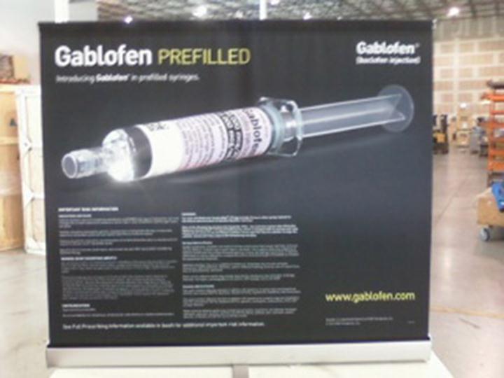 Of a black banner with the text "Gablofen Prefilled" and an image of a syringe. The banner is displayed in a warehouse or storage area with shelves and boxes in the background. The syringe is shown in the center of the banner with the needle pointing towards the right side of the image. The text on the banner is in white and black and it appears to be an advertisement for the product. The website "www.gablfen.com" is also visible on the bottom right corner.