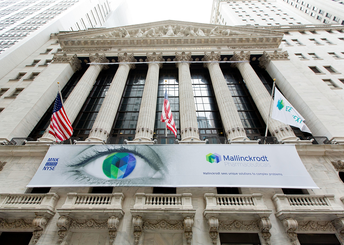 The exterior of the New York Stock Exchange building in New York City. The building is a neoclassical structure with columns and intricate carvings on the facade. The entrance of the building is flanked by two American flags on either side. In the center of the image there is a large banner with the words "Mallinckrodt" and an image of an eye. The banner also has the logo of the company. The sky is overcast and there are other skyscrapers visible in the background.