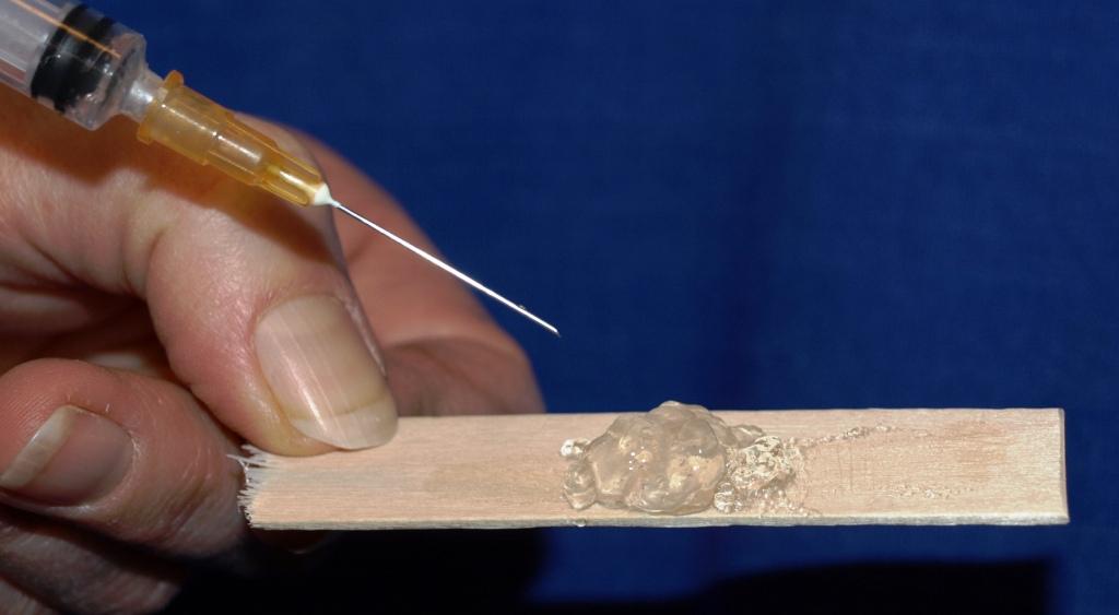 A person's hand holding a syringe. Its needle is pointing towards a small piece of wood. On the wood there is a small amount of a clear substance a thumbnail-sized amount of what appears to be a clear gel. The background is a solid blue color.