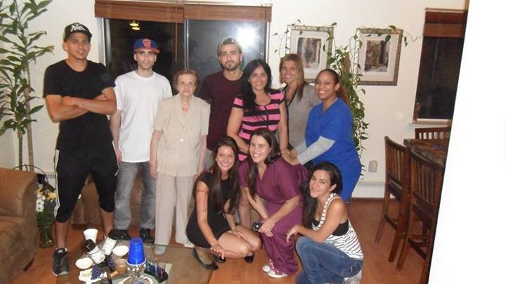 A group of ten people posing for a photo in a living room. All are smiling and looking at the camera. The group is arranged in a semi-circle. Seven people are standing and three women are squatting in front. <br /><br />In the center of the group there is an older woman there is a coffee table with beer bottles and Solo cups on it. The room is decorated with plants and a painting hanging on the wall.