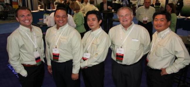 A group of five men standing together in a conference room. They are all wearing white shirts and black pants and have red lanyards around their necks. The men are smiling and appear to be posing for the photo. In the background there are other people and tables with various items on them. The room appears to be well-lit with blue carpet and large windows.