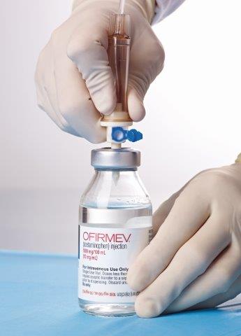 A person's hand wearing a white glove holding a syringe and injecting a vaccine into a small glass vial. The vial is labeled "OFIRMEV" and has a blue cap. The background is blurred but it appears to be a laboratory setting with a blue tablecloth. The person's hands are positioned in a way that suggests they are about to administer the vaccine.