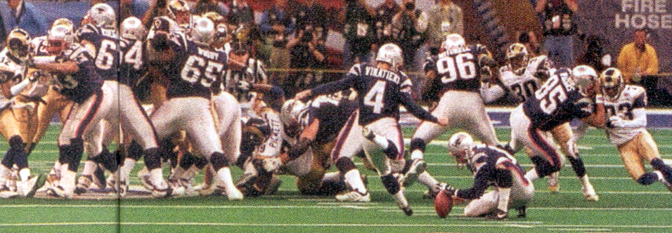 A photograph of a football game in progress. It shows a group of players from both teams on the field during a game. The players are wearing white and gold uniforms with numbers on their backs and helmets. The player in the center of the image number 4 is diving into the end zone for a touchdown. He is holding the ball and is being tackled by the opposing team number 96 who is wearing a black and gold uniform. The other players are surrounding him trying to tackle him. The field is covered in green turf and there are spectators in the background. The image appears to have been taken from a high angle looking down on the action.