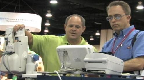 Two men standing in front of a table with a medical equipment on it. The man on the left is wearing a green t-shirt and is holding a white machine with a blue handle. He appears to be explaining something to the other man who is also wearing a blue shirt and glasses. The table is covered with a white tablecloth and there is a sign in the background that reads "Medical Equipment Show". The men appear to be at a trade show or exhibition.