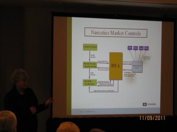 A woman standing in front of a large screen with a presentation on it. The presentation is titled "Narcotics Market Controls" and it appears to be a flowchart or diagram. The flowchart is divided into different sections each representing a different aspect of the Narcotics market. <br /><br />The first section is labeled "DEA" and shows the different components of the market. The second section is labeled "Product Development" and has a list of products and services. The third section has a description of the products such as drugs pharmaceuticals and pharmaceuticals. The fourth section has an image of a person's face and the date "11/09/2011" is displayed at the bottom right corner of the screen.<br /><br />There are people sitting in the audience attentively listening to the presentation. The background is dark and the overall mood of the image is professional and focused.