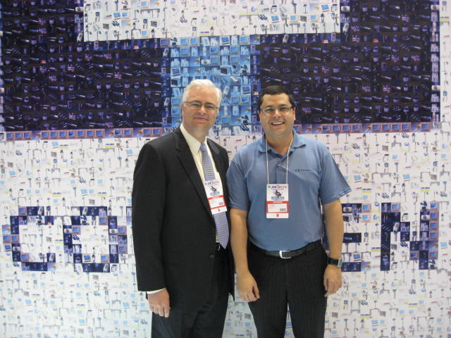 Two men standing side by side in front of a large wall covered in blue and white mosaic tiles. The tiles are arranged in a grid-like pattern creating a mosaic-like effect. The man on the left is wearing a black suit and tie while the man in the blue shirt is wearing glasses and a name tag. Both men are smiling and appear to be posing for the photo. The background is a white wall with a blue border.