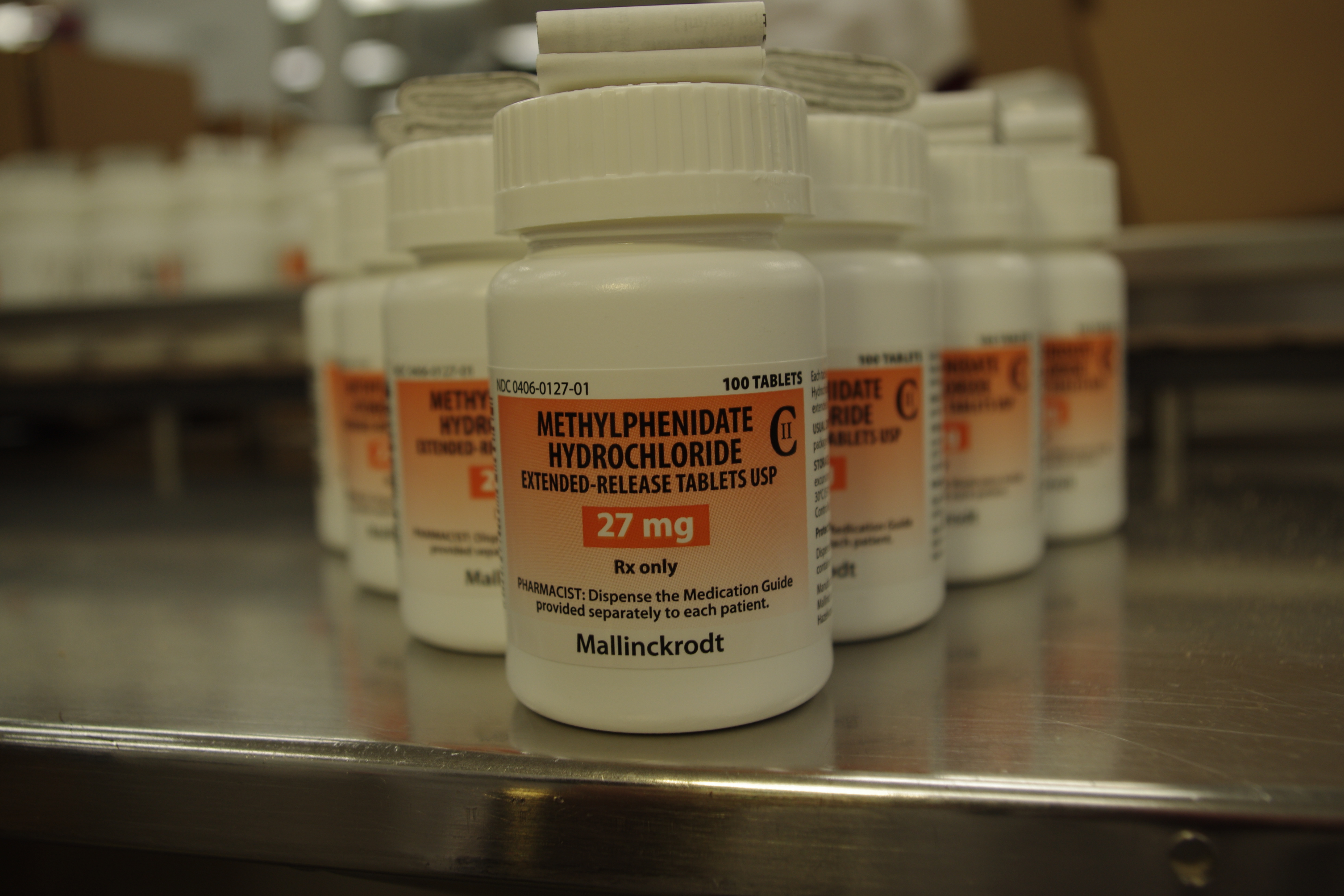 A row of white medicine bottles with orange labels on them. The bottles are arranged in a neat row on a metal shelf. The label on the bottles reads "Methylphenidate Hydrochloride 27 mg" and "Mallinckrodt". The bottles appear to be new and unused. The background is blurred but it seems to be a warehouse or storage area with other bottles and boxes visible.