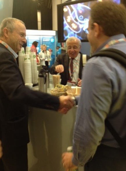 Two men shaking hands in an event. The man on the left is wearing a black suit and is smiling widely. He appears to be in his late 60s or early 70s. He is standing in front of a counter with a tray of food and a coffee machine on it. On the right side of the image there is another man wearing a blue shirt and carrying a backpack. In the background there are other people and a large screen displaying an image of a fish.