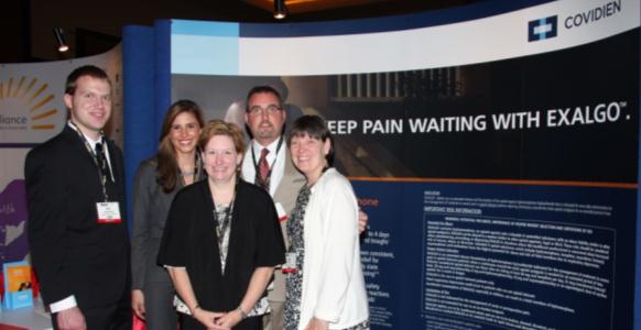 A group of five people standing in front of a blue and white banner with the text "Keep pain waiting with Exalgo" and the logo of Covidien. The banner is located at an exhibition or trade show booth. The people in the photo are smiling and appear to be posing for a photo. On the left side of the image there is a man wearing a suit and tie and on the right side there are two women and a man. The background is dark and there are other banners visible in the background.