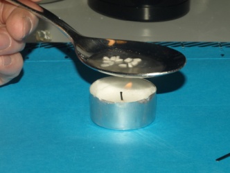 A hand holding a silver spoon over a small white candle. The candle is placed on a blue surface with a black stovetop in the background. The spoon is resting on top of the candle with the handle of the spoon slightly tilted to the side. The handle is covered in small white flowers which appear to be floating in the air. The background is blurred but it appears to be a kitchen countertop.