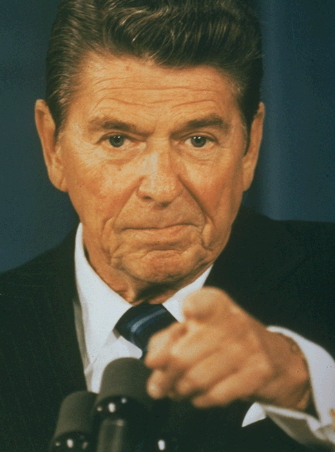 A close-up portrait of former US President Ronald Reagan. He is wearing a dark suit and tie and is sitting at a table with microphones in front of him. He appears to be speaking into one of the microphones as he is gesturing with his right hand as if he is making a point. His expression is serious and he is looking directly at the camera. The background is blurred so the focus is on Reagan's face.