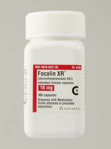 Of a white plastic bottle with a white cap. The label on the bottle is red and white with black text. The text reads "Focalin XR" and "10 mg". Below the text there is a description of the product which states that it is an extended-release capsule with 100 capsules. The bottle also mentions that the product is a medication guide attached or provided separately. The brand name "Novartis" is written in black text at the bottom of the label.