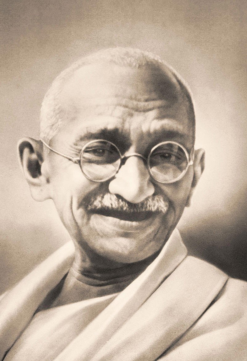 A black and white portrait of Mahatma Gandhi the founder of India. He is shown from the waist up looking directly at the camera with a slight smile on his face. He has a mustache and is wearing round glasses with thin frames. His hair is neatly combed back and he is wearing a traditional Indian dhoti. The background is blurred making the focus of the image the focal point. The image appears to be a close-up of Gandhi's face and upper body.