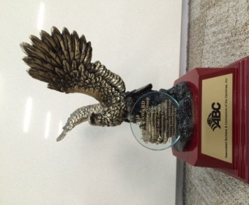 A trophy or award in the shape of a bird. The trophy is made of metal and has a red base with the letters "ABC" on it. The bird is depicted in flight with its wings spread wide and its head turned to the side. The wings are intricately detailed and the body of the bird is covered in small beads. The beads are arranged in a circular pattern around the bird's head and neck. The background is a beige carpeted floor.