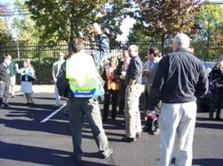 A group of people gathered in a parking lot. There are several people in the group some of whom are standing and some are walking. In the center of the image there is a man wearing a yellow vest and carrying a blue backpack. He appears to be a police officer or a member of the public. He is standing with his arms raised in the air as if he is directing the group. Behind him there are trees and a fence. The sky is blue and there are a few clouds in the sky. It seems like the group is participating in a protest or demonstration.