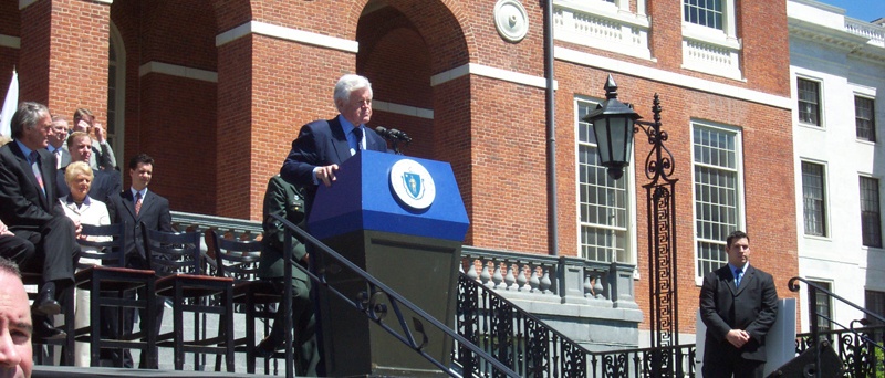 A man standing at a podium in front of a large brick building. The man is wearing a suit and tie and appears to be giving a speech or presentation. He is speaking into a microphone and is standing on a balcony with a railing. Behind him there are several people sitting on chairs some of whom are looking at him attentively. On the right side of the image there is a man in a suit standing on the balcony. The building behind the man is white with columns and a clock on the wall. The sky is blue and there are trees in the background.