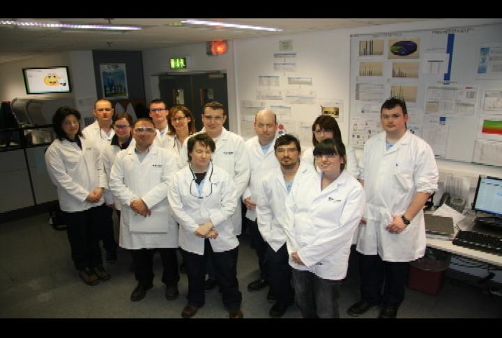 A group of nine people all wearing white lab coats standing in a laboratory. They are all looking at the camera and appear to be posing for a group photo. The laboratory has a whiteboard on the right side of the image with various scientific posters and charts pinned to it. There is a computer monitor and keyboard on the desk in the background. The people in the photo are of different ages and ethnicities and they are all smiling and looking towards the camera.