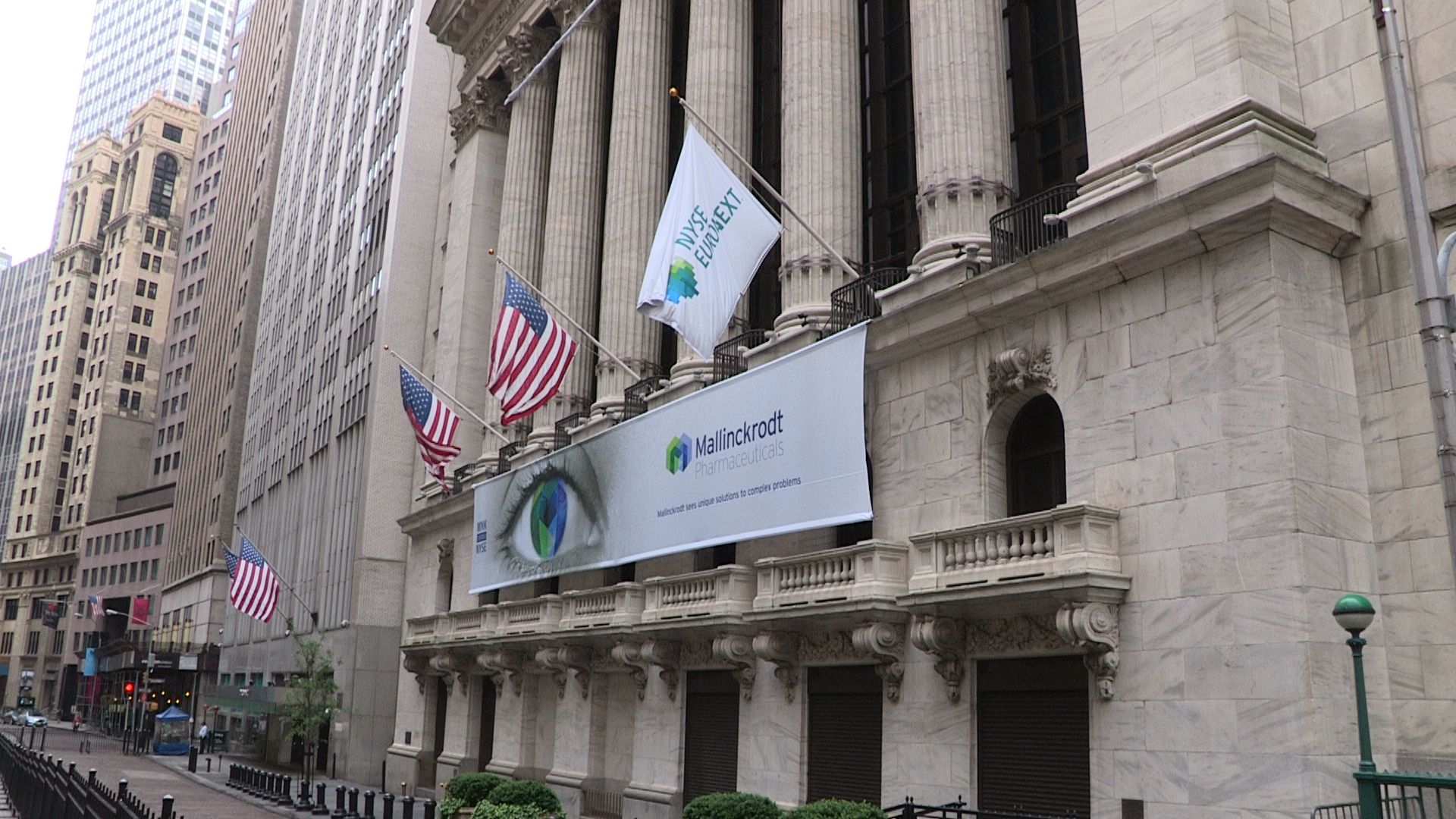 A large beige-colored building with columns on the side of it. The building appears to be a bank or a financial institution as there is a large banner hanging from the side with the logo of the bank and the words "Bank of America" on it. There are several American flags flying in front of the building including the American flag and the United States flag. The street is lined with other tall buildings and there are people walking on the sidewalk. The sky is overcast and the overall mood of the image is gloomy.