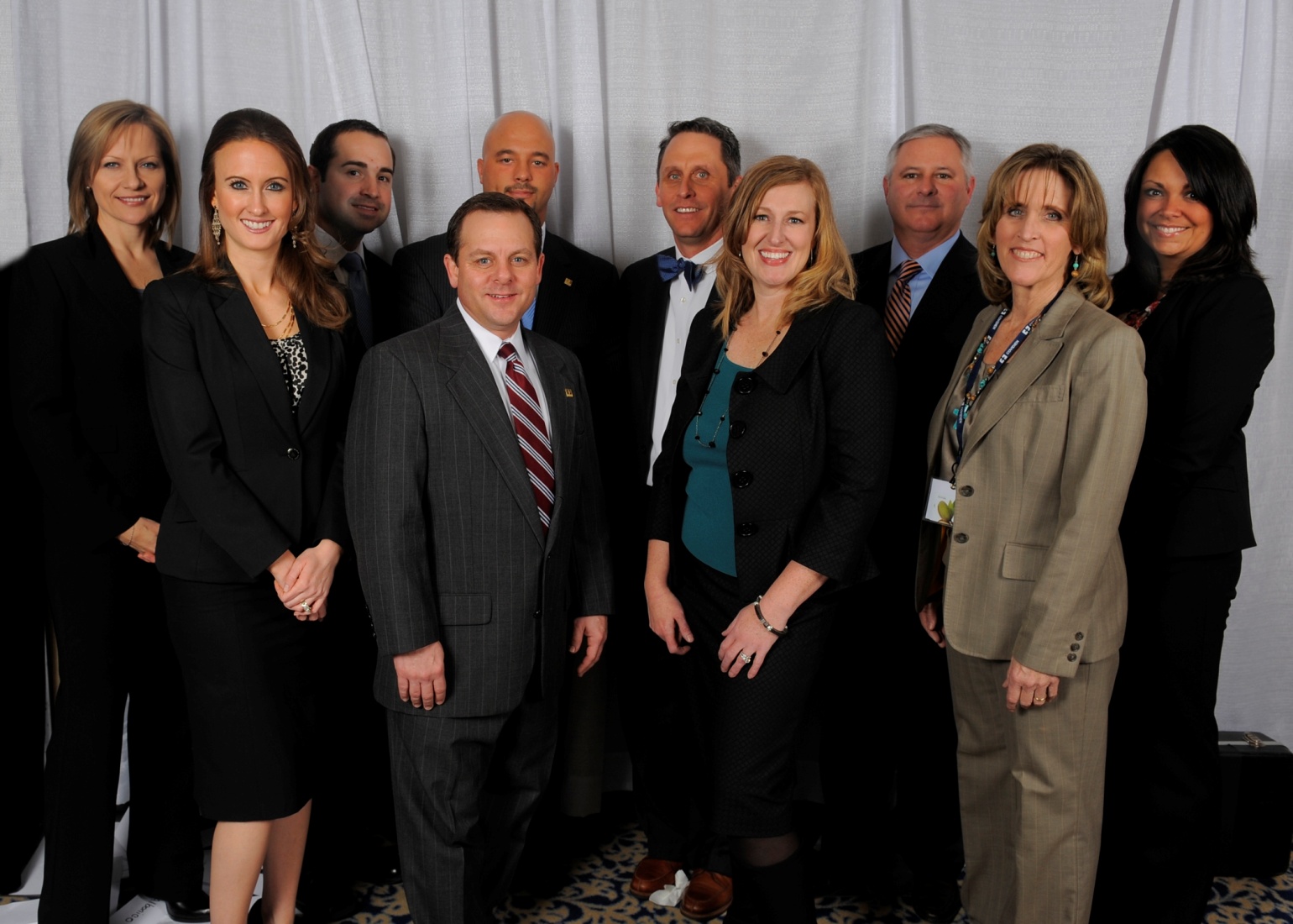 A group of ten people all dressed in formal business attire. They are standing in front of a white curtain and are facing the camera with happy expressions.