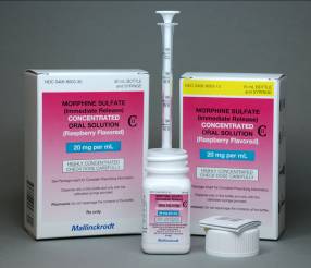 A set of medical supplies including a syringe a box and a small bottle. The syringe is white with a pink label that reads "Morphine Sulfate Concentrated Oral Solution (Raspberry Flavored)". The box is white and has a pink and yellow label with the product name and description. The bottle is also white and is placed next to the syringe. There is also a small white cap on the right side of the image. The background is a plain grey color.