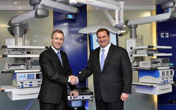 Two men shaking hands in front of a medical equipment set up in a hospital room. The men are both wearing suits and are smiling at the camera. Behind them there are several pieces of medical equipment including a CT scanner machine. The room has blue walls and a large window on the left side allowing natural light to enter. On the right side there is a sign that reads "Covidien". The men appear to be in a professional setting likely in a medical facility.