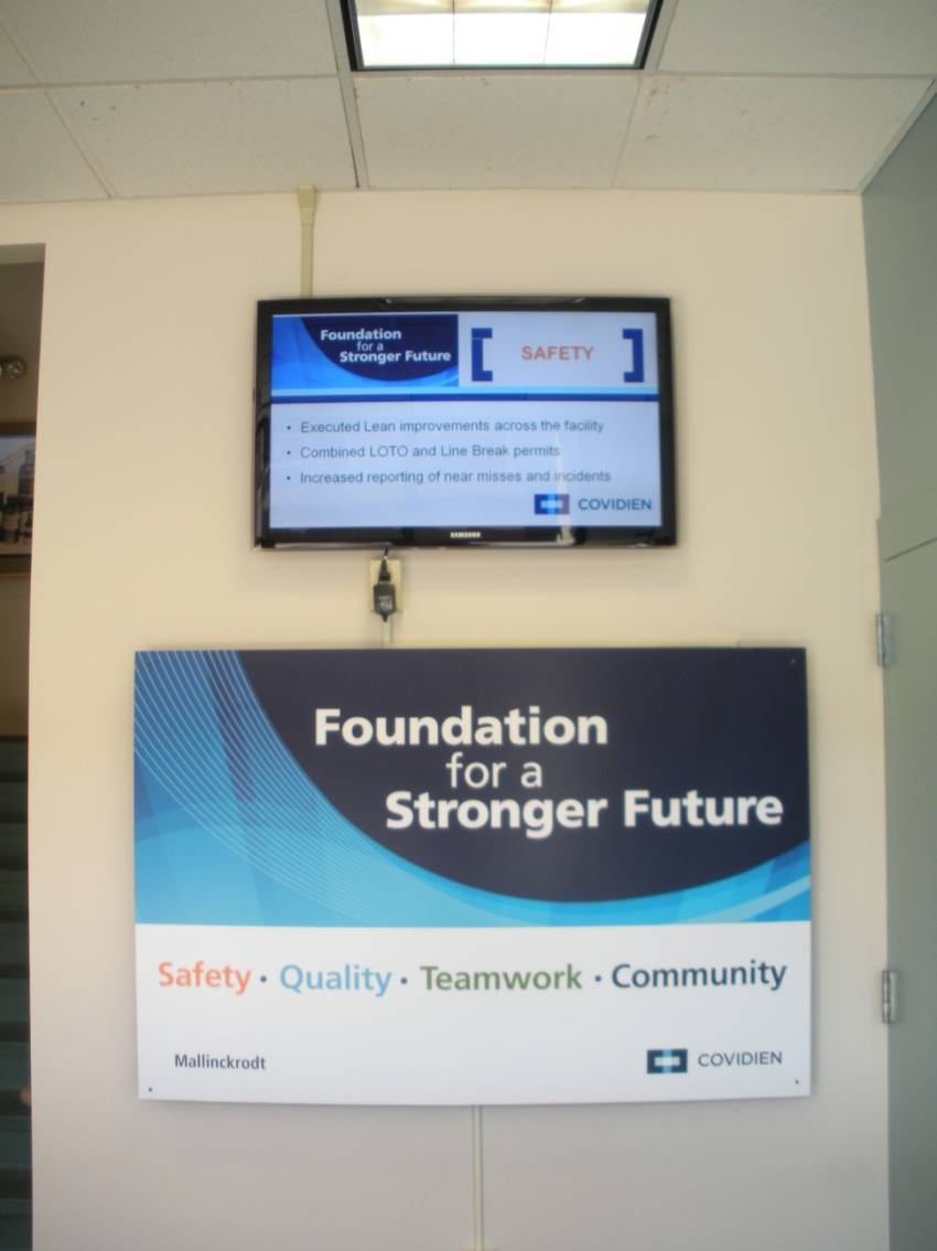 A large blue and white sign hanging on a white wall in a hallway. The sign reads "Foundation for a Stronger Future" in bold white letters. Below the sign there is a smaller sign that reads "Safety - Quality - Teamwork - Community" in smaller white letters with the logo of Covidien on the bottom right corner. Above the sign is a flat-screen TV mounted on the wall. The TV screen displays a message about the foundation for a stronger future. The hallway appears to be well-lit with natural light coming in from the windows on the left side of the image.