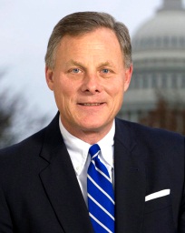 A portrait of a middle-aged man in a suit and tie. He is standing in front of the United States Capitol building which is visible in the background. The man is smiling and looking directly at the camera. He has short light-colored hair and is wearing a dark suit with a white shirt and a blue striped tie. The image appears to be a professional headshot.