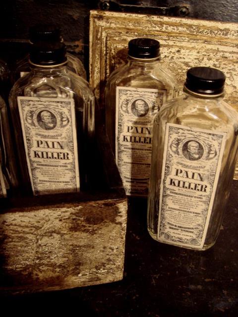 Three glass bottles with black lids and labels on them. The labels have the words "Pain Killer" written in bold black letters. The bottles are arranged in a row with one bottle on the left one in the middle and one on the right. The background is dark and there is a wooden box on the floor next to the bottles. The box appears to be old and weathered with peeling paint and rust visible on the surface. The overall mood of the image is eerie and mysterious.