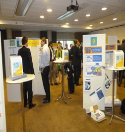 A group of people gathered in a large room with a high ceiling and a projector screen. There are several white banners with blue and yellow text and images on them as well as a table with a sign that reads "13". The people in the image appear to be engaged in a discussion with some standing and others walking around. The room appears to be a conference or exhibition space as there are other people visible in the background.