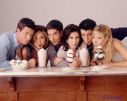 A publicity photograph for the tv show Friends. It shows the six main characters sitting at a wooden table with milkshakes and ice cream sundaes in front of them. The two milkshakes each have three straws and all the characters are drinking from one of the straws. The background is a plain white wall. The overall mood of the image is casual and relaxed.