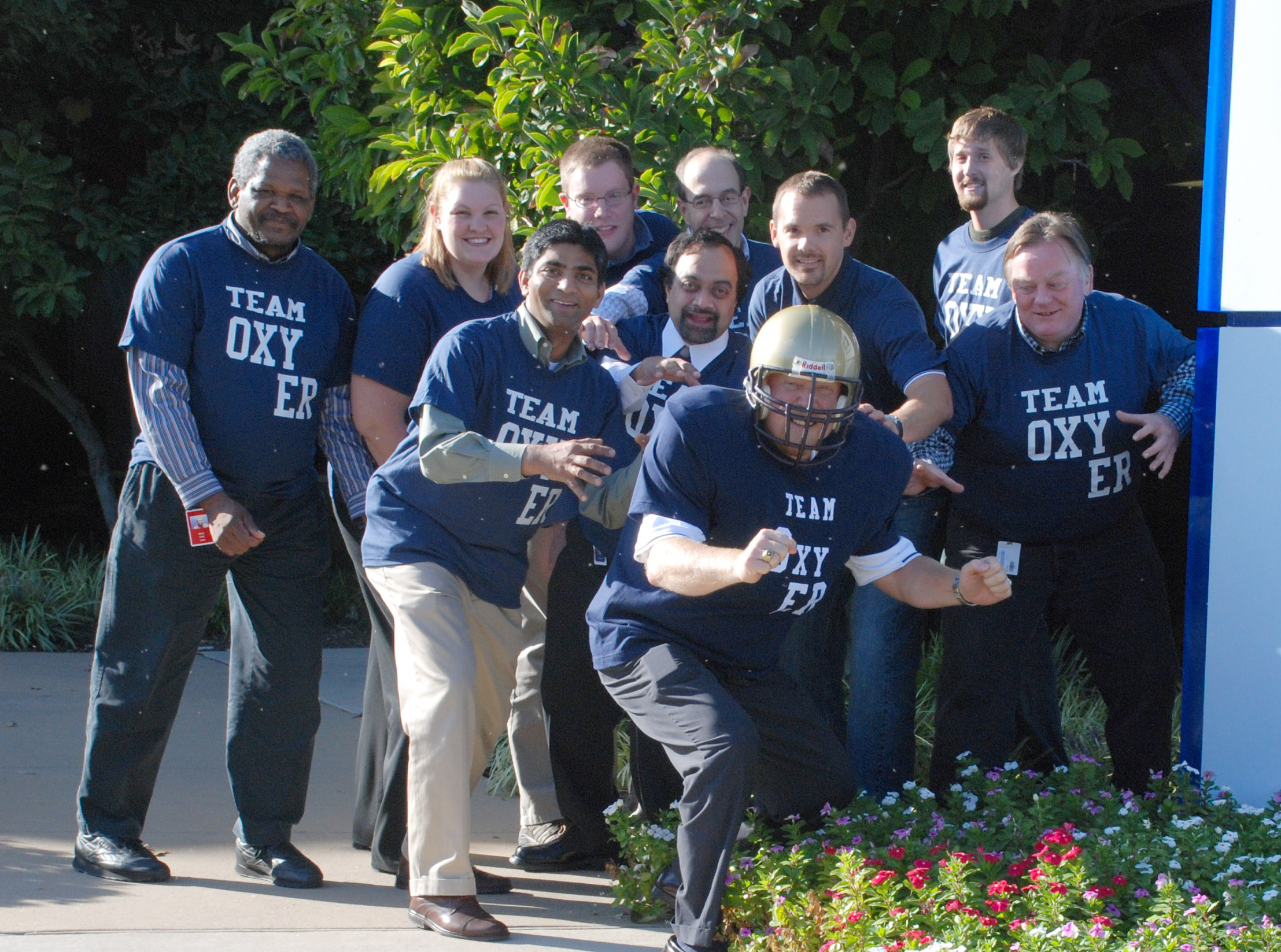 A group of nine people all wearing blue t-shirts with the words "Team Oxygen" printed on them. They are standing in front of a blue and white sign that reads "Oxygen" and are posing for a photo. In the center of the group there is a man wearing a football helmet and holding a football. He is in a crouching position with his arms stretched out to the sides and a big smile on his face. Behind him there are trees and bushes with colorful flowers. The group appears to be posing for the photo.