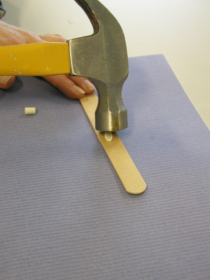 A person's hand holding a yellow hammer. The hammer is aimed toward a small yellow pill sitting on top of a wooden tongue depressor. The tongue depressor is sitting on a purple mat. There is also a small white object possibly another pill on the surface next to the hammer.