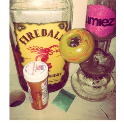 A collection of items on a table. From left to right : A bottle of Fireball Cinnamon Whiskey  a prescription pill bottle (from Rite Aid) and a bong (with a pink sticker from Zumiez an action sports clothing store chain). There is also a small piece of green paper in the foreground.