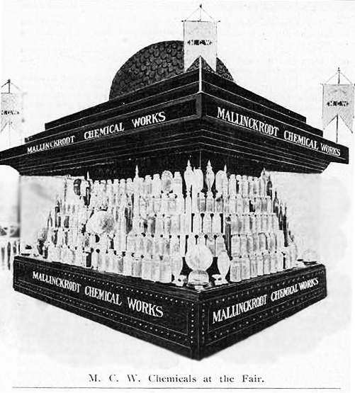 A black and white photograph of a square display booth at a fair. The image appears to be color reversed. The display is made up of multiple rows of glass bottles arranged in a pyramid-like structure. The bottles are of different sizes and shapes. The top of the display has a dome-shaped structure.  Above and below the bottles are signs that read: "Mallinckrodt Chemical Works" in bold letters. The image is labeled "M. C. W. Chemicals at the Fair." The image appears to date to the early twentieth century.