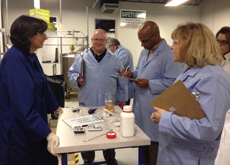 A group of people in a laboratory setting. There are six people in the image all wearing blue lab coats. They are gathered around a table with various scientific equipment and tools on it. The person in the center of the image is holding a clipboard and appears to be explaining something to the group. To his left there is a woman wearing a blue lab coat and gloves and to his right there are two men wearing lab coats and glasses. Behind them two men are standing and looking at the table. In the background we can see a safety sign and other equipment.