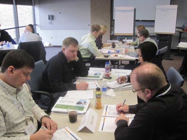 A group of people sitting around a long table in a conference room. They are all holding pens and appear to be engaged in a discussion. The table is covered with papers books and water bottles and there is a whiteboard in the background. The room has large windows on the left side allowing natural light to enter. The people are dressed in casual business attire and are focused on their work.