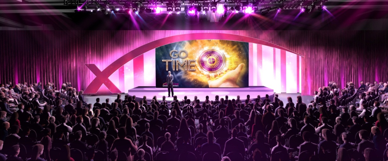 A large stage with a large screen in the center. The screen displays the words "Go Time" in bold colorful letters. The stage is surrounded by a pink curtain and there are rows of people sitting in front of it facing the stage. The audience is seated in rows of chairs and the stage is lit up with purple and pink lights. The overall atmosphere of the image is one of excitement and anticipation.