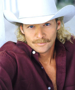 A close-up portrait of a man wearing a white cowboy hat. He has blonde hair and a mustache and is wearing a maroon button-down shirt. He is looking directly at the camera with a slight smile on his face. The background is blurred but it appears to be an outdoor setting with a white wall and a window. The man's expression is confident and relaxed.