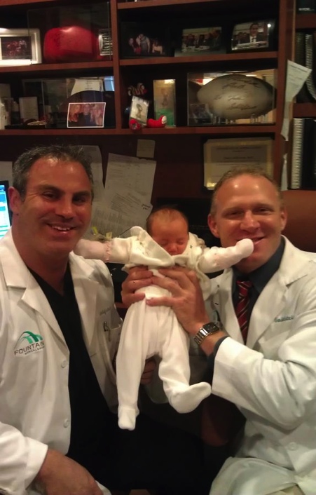 Two men in white lab coats holding a newborn baby. The man on the left is holding the baby in his arms and is smiling at the camera. The baby is wearing a white onesie and is lying on its back with its head resting on its hands. The other man is sitting next to the baby and is also smiling. In the background there is a bookshelf with various medical supplies and a computer monitor. Both men appear to be happy and proud of the baby.