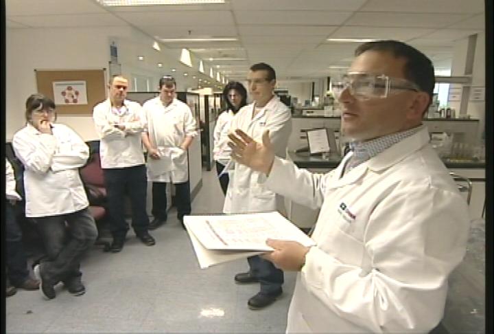 A group of six people in a laboratory setting. They are all wearing white lab coats and are standing in a line facing the camera. The person in the center of the image is a man wearing a white lab coat and safety goggles and he is holding a clipboard and appears to be explaining something to the group. He is gesturing with his hands as he speaks. Behind him there are several other people some of whom are also wearing lab coats and they are looking at him attentively. The room is well-lit with fluorescent lights and there are various scientific equipment and equipment visible in the background.