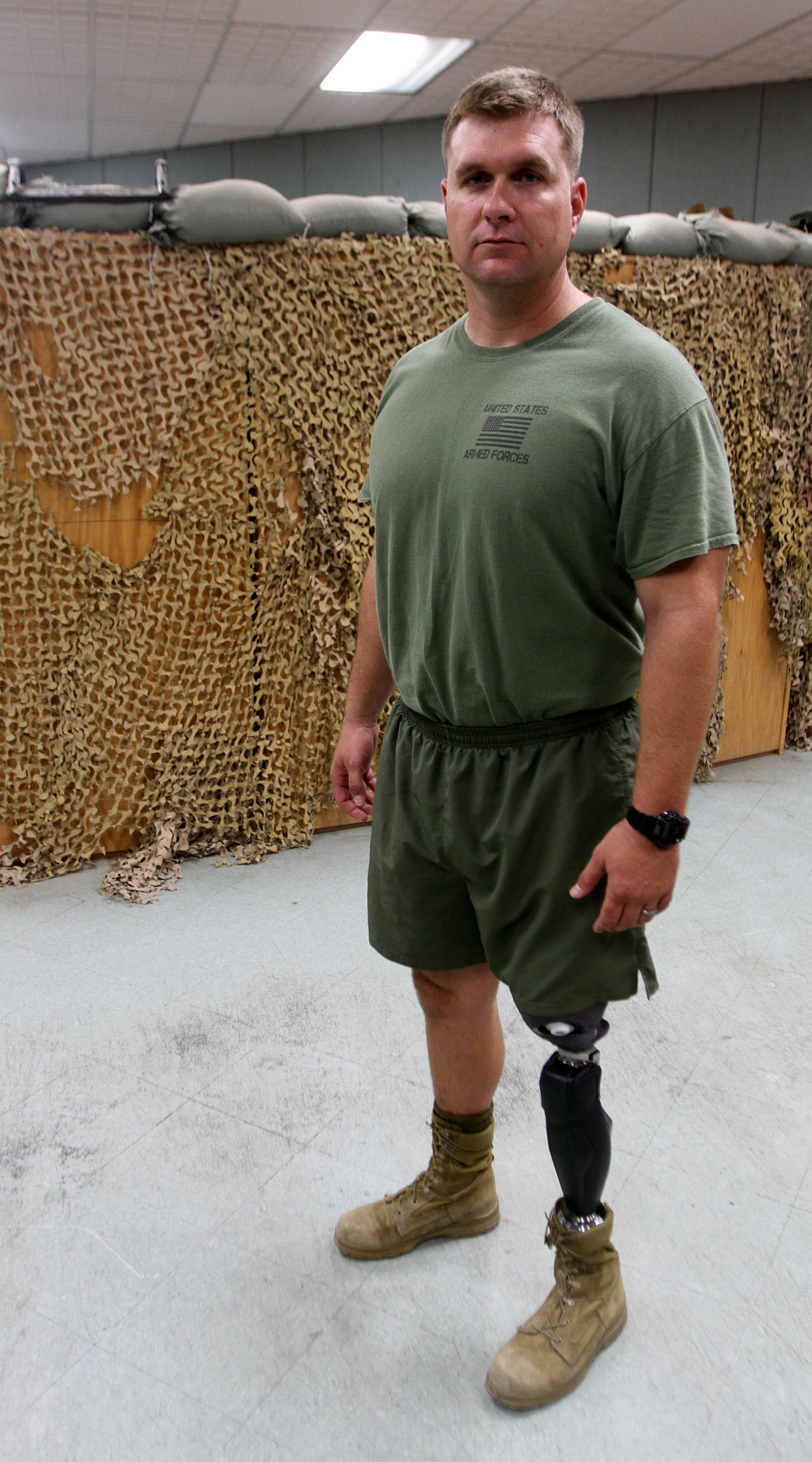 A man standing in a room with a camouflage wall in the background. He is wearing a green t-shirt green shorts and beige boots. He has a black prosthetic leg and is looking directly at the camera with a serious expression on his face. The man appears to be in his late twenties or early thirties.